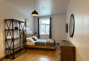 A bedroom with a bed, a wooden dresser, a black metal shelf unit, a hanging light fixture, a window with curtains, and a wall-mounted circular object.