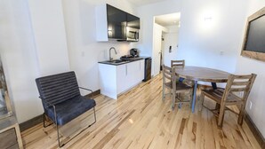 Et kompakt køkken med mikrobølgeovn, vask og skabe. Der er et spisebord med fire stole. En moderne stol er placeret nær køkkenet.
