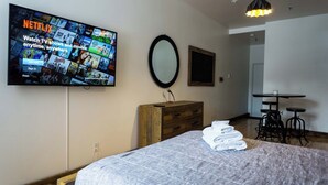 A bedroom with a bed, a wooden dresser, a TV mounted on the wall displaying Netflix, a mirror, and a small table with stools.