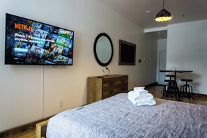 A bedroom with a bed, a wooden dresser, a TV mounted on the wall displaying Netflix, a mirror, and a small table with stools.
