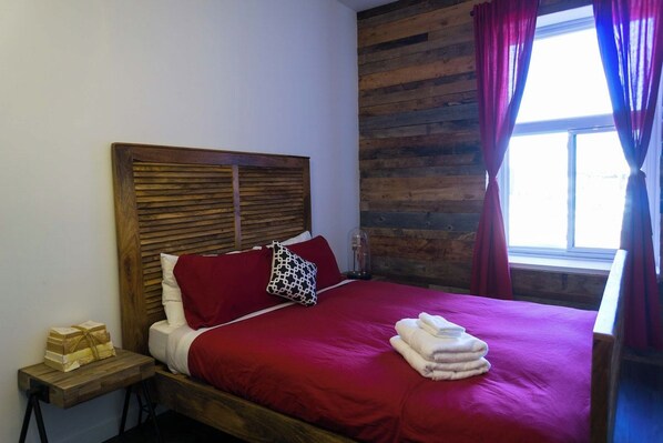 A bed with a wooden headboard, red bedding, and a window with curtains.