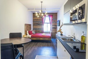 A compact room with a bed, a dining table, a kitchen area, and a window with curtains.