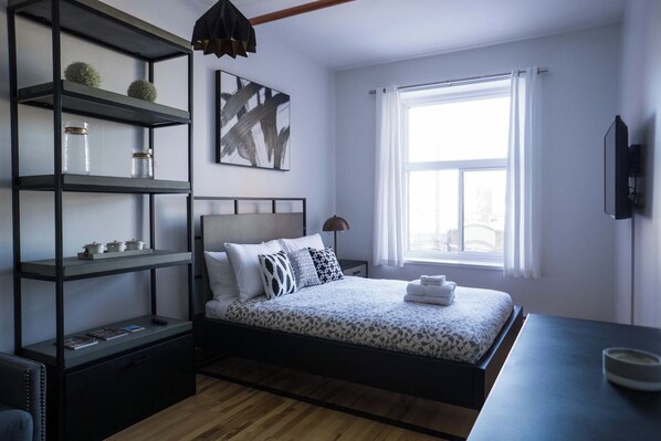 A bedroom with a bed, a nightstand, a shelf unit, a painting, a window, and a television.
