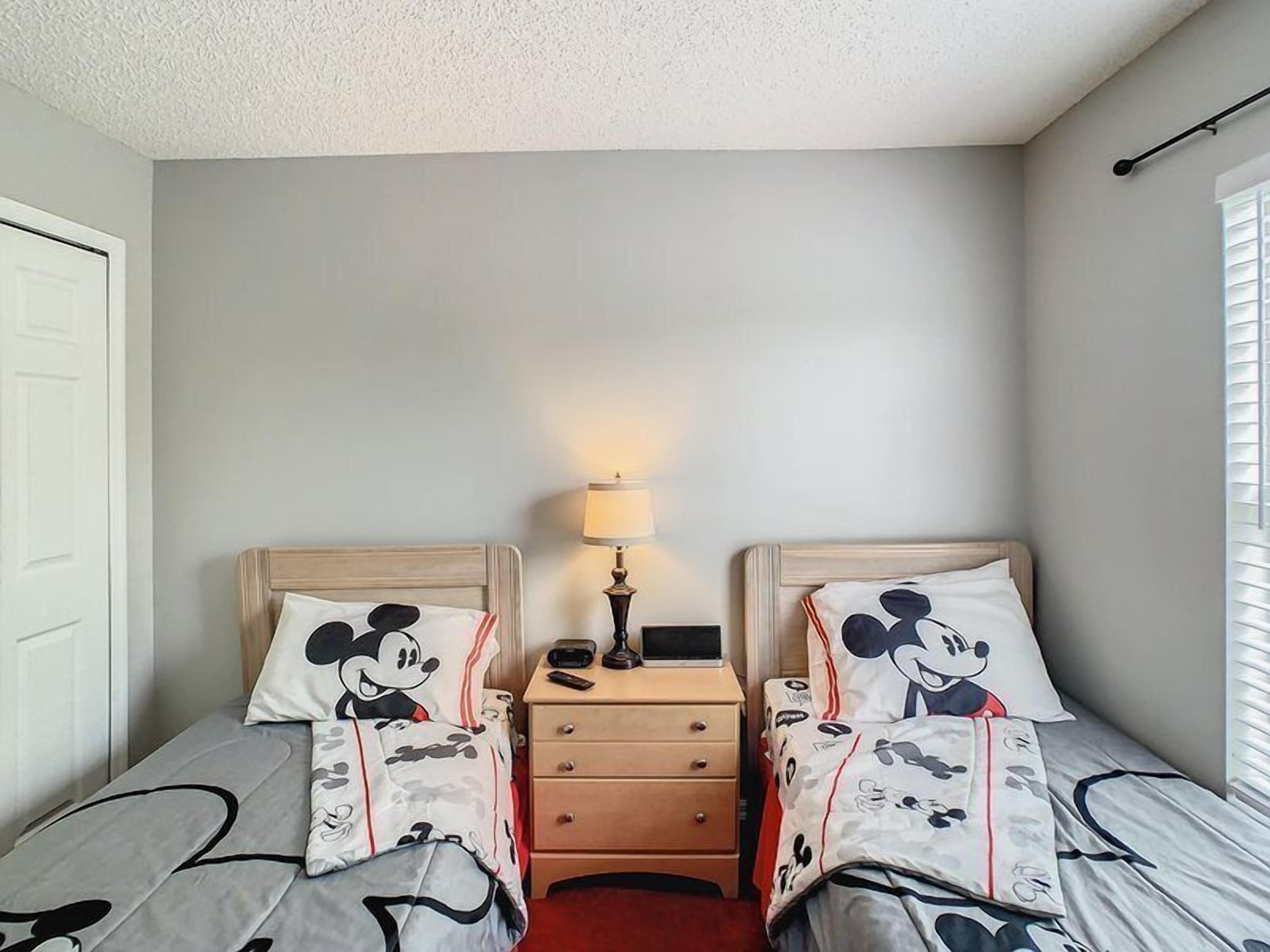Habitación de hotel con dos camas, cada una con ropa de cama temática de Mickey Mouse. Hay una mesita de noche con una lámpara y un control remoto.