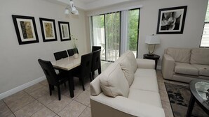 Un salon avec un canapé beige, une table à manger avec des chaises et une table basse. Les murs sont ornés de tableaux encadrés et il y a une fenêtre avec des stores.
