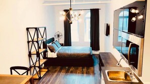 A compact bedroom with a bed, a desk, a chair, a small table, a built-in sink, and a large mirror.