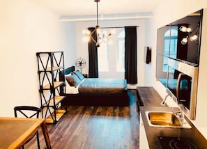 A compact bedroom with a bed, a desk, a chair, a small table, a built-in sink, and a large mirror.