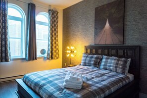 Une chambre à coucher comprenant un lit, une tête de lit, une œuvre d’art accrochée au mur, une fenêtre avec des rideaux et une lampe sur pied.