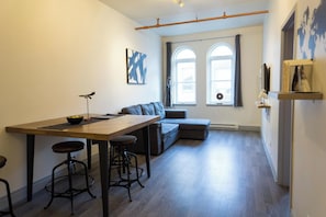 Un espace de vie moderne comprenant une table à manger en bois, deux tabourets de bar, un canapé gris et une grande fenêtre avec des rideaux.