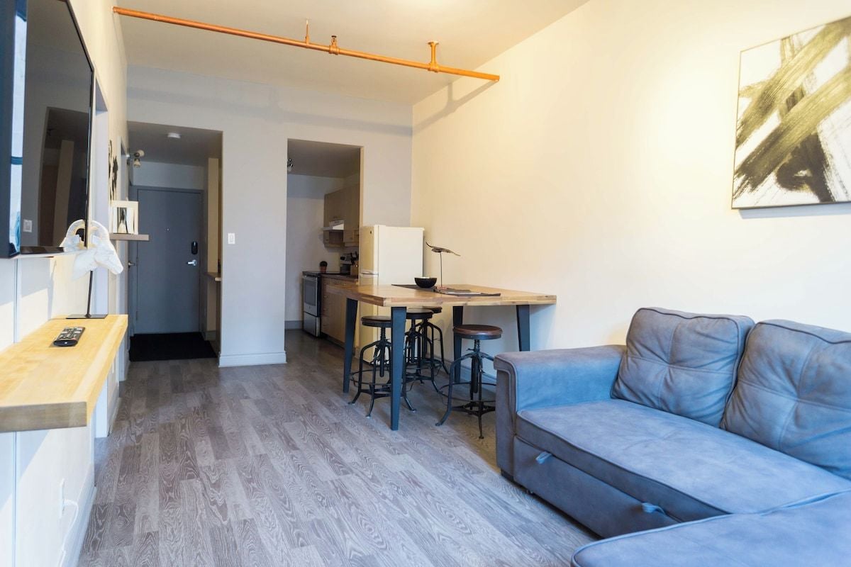 A modern living space with a sofa, a dining table with stools, a kitchen area, and a hallway leading to another room.