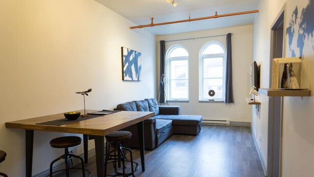 En moderne stue med et træbord, to barstole, en grå sofa og et stort vindue med gardiner.
