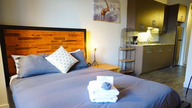 A hotel room with a wooden headboard, a bed with blue bedding, a nightstand with a lamp, and a kitchenette with stainless steel appliances. - Intimate loft in the heart of the city (Québec)