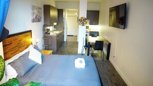 A hotel room with a bed, a desk, a chair, a small kitchen area, and a dartboard.