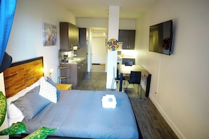 A hotel room with a bed, a desk, a chair, a small kitchen area, and a dartboard.