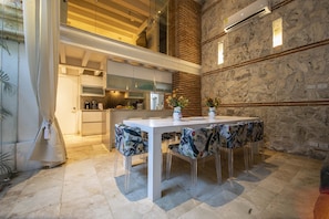 Une cuisine moderne avec un plafond haut, un coin repas avec une table blanche et des chaises à motifs, et un mur en pierre doté d’un éclairage intégré.