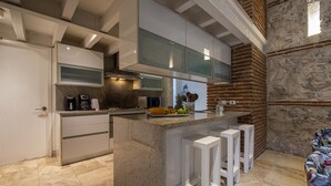 A modern kitchen with a central island, white bar stools, and a brick accent wall. - Santos de Piedra Exclusive Home (Provincia de Cartagena)