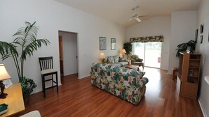 Ein geräumiges Wohnzimmer mit Holzboden, einem floralen Sofa, einem Esstisch mit Stühlen und einem Deckenventilator.