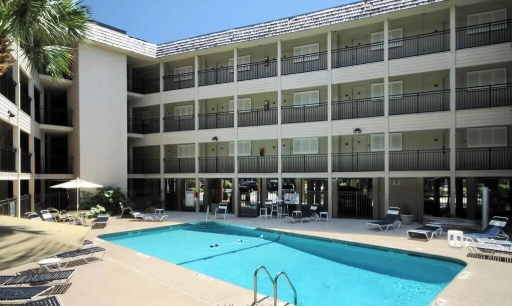A multi-story hotel with a swimming pool, outdoor seating, and a palm tree.