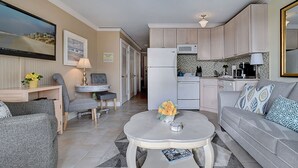 A modern living room with a grey sofa, a coffee table with a vase of flowers, a TV mounted on the wall, and a kitchen area with light-colored cabinets and appliances. - Awesome Condo! King size bed! Updated! Ocean Views! 2 Bikes! #218 Great Reviews! (Hilton Head Island)