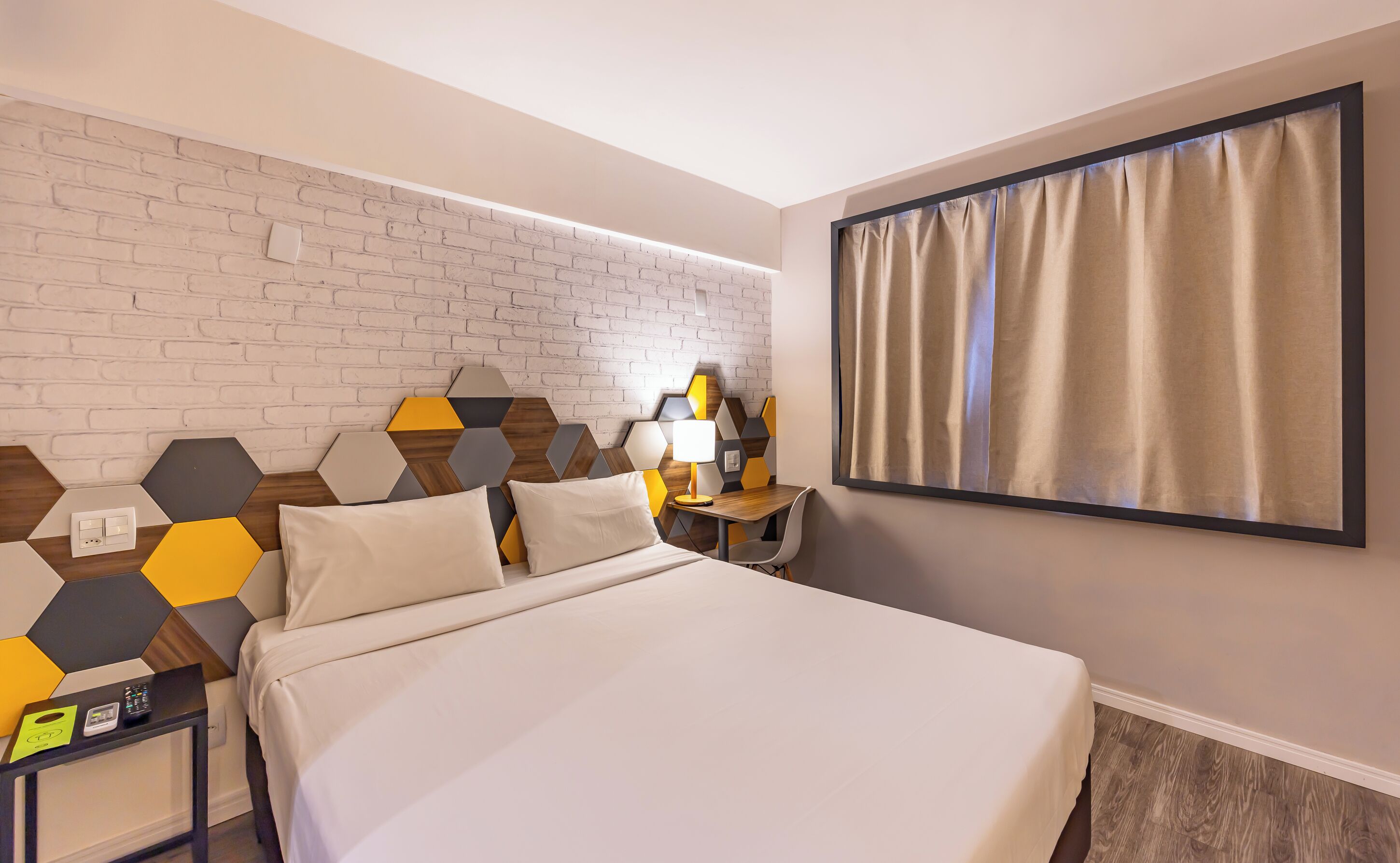 Ein Hotelzimmer mit einem Bett, einem Schreibtisch mit Lampe, einer Ziegelwand und einem mit Vorhängen drapierten Fenster.
