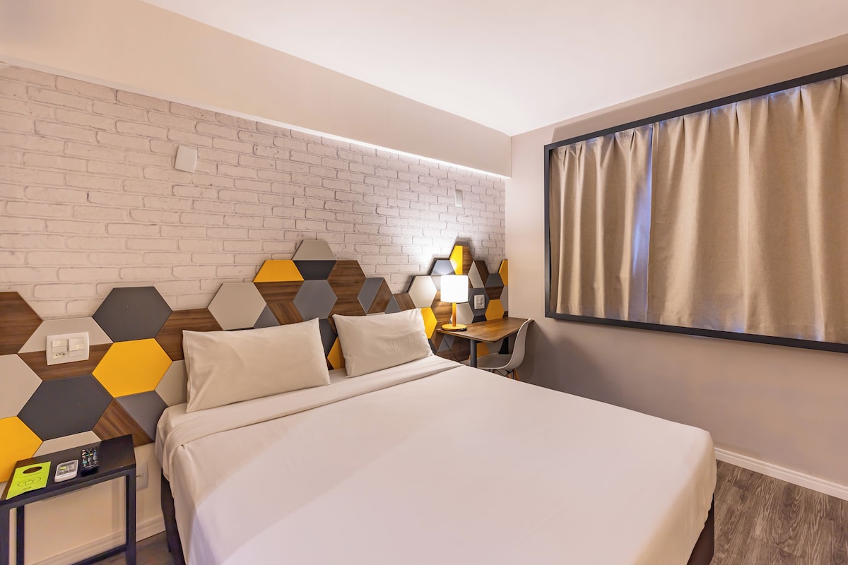 A hotel room with a bed, a desk with a lamp, a wall with a brick pattern, and a window with curtains.