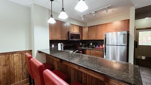 A modern kitchen with wooden cabinets, a stainless steel refrigerator, a microwave, and a granite countertop. - Charming Fire Mountain Lodge Condo | Walk to Downtown | Sleeps Six  (Canmore)