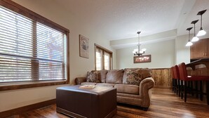 A living room with a brown leather sofa, a wooden dining table with red chairs, and a large window with blinds. - Charming Fire Mountain Lodge Condo | Walk to Downtown | Sleeps Six  (Canmore)