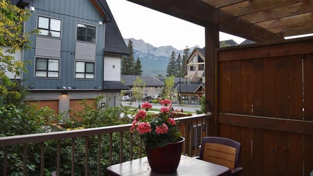 A balcony with a wooden railing overlooking a mountain view, a table with a potted plant, and a chair. - Charming Fire Mountain Lodge Condo | Walk to Downtown | Sleeps Six  (Canmore)