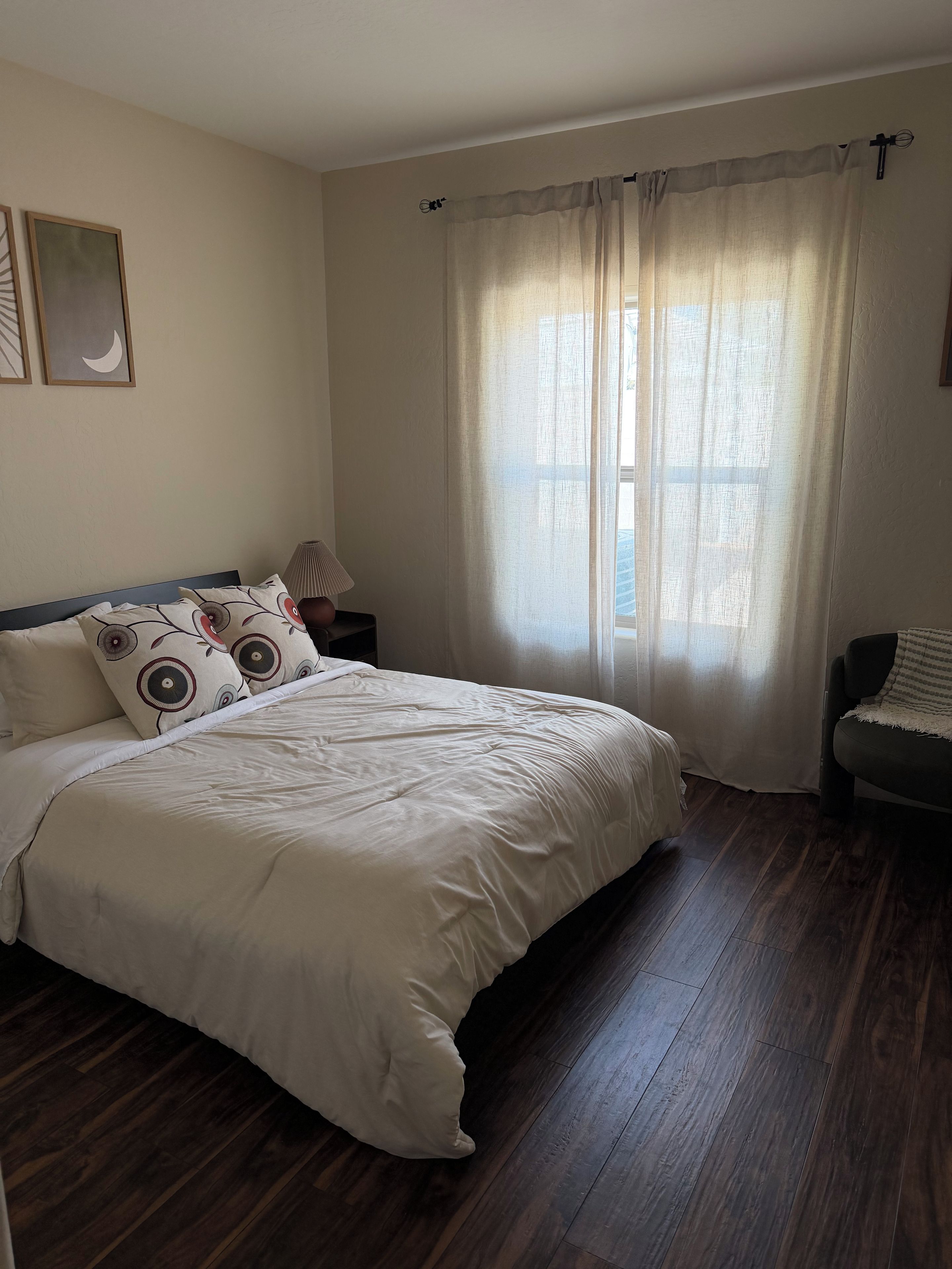A bedroom with a bed, a nightstand, a lamp, a chair, and a window with curtains.