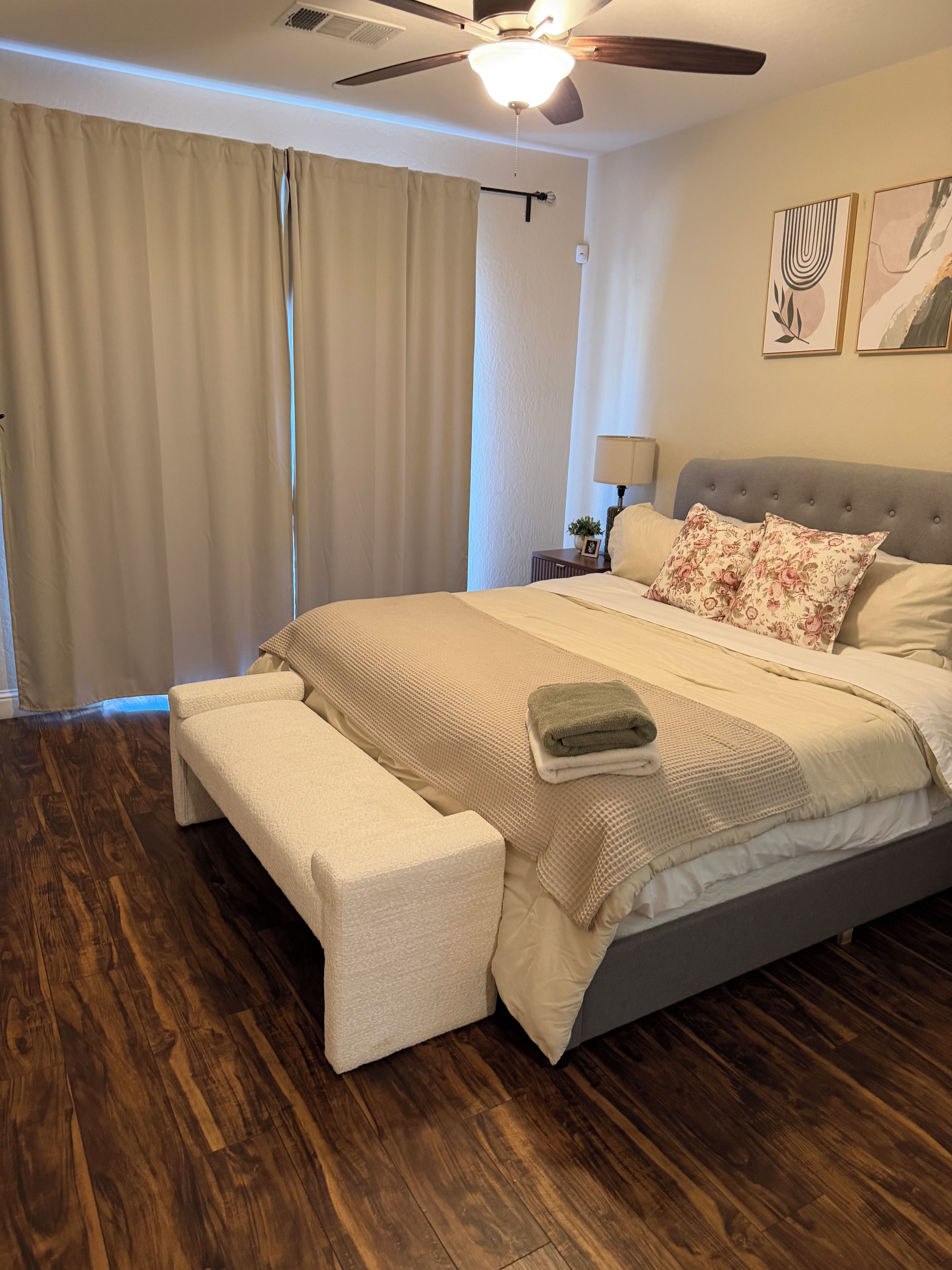 Quarto com cama, um banco, ventilador de teto e janela com cortinas.