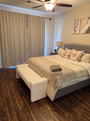 Een slaapkamer met een bed, een bankje, een plafondventilator en een raam met gordijnen.