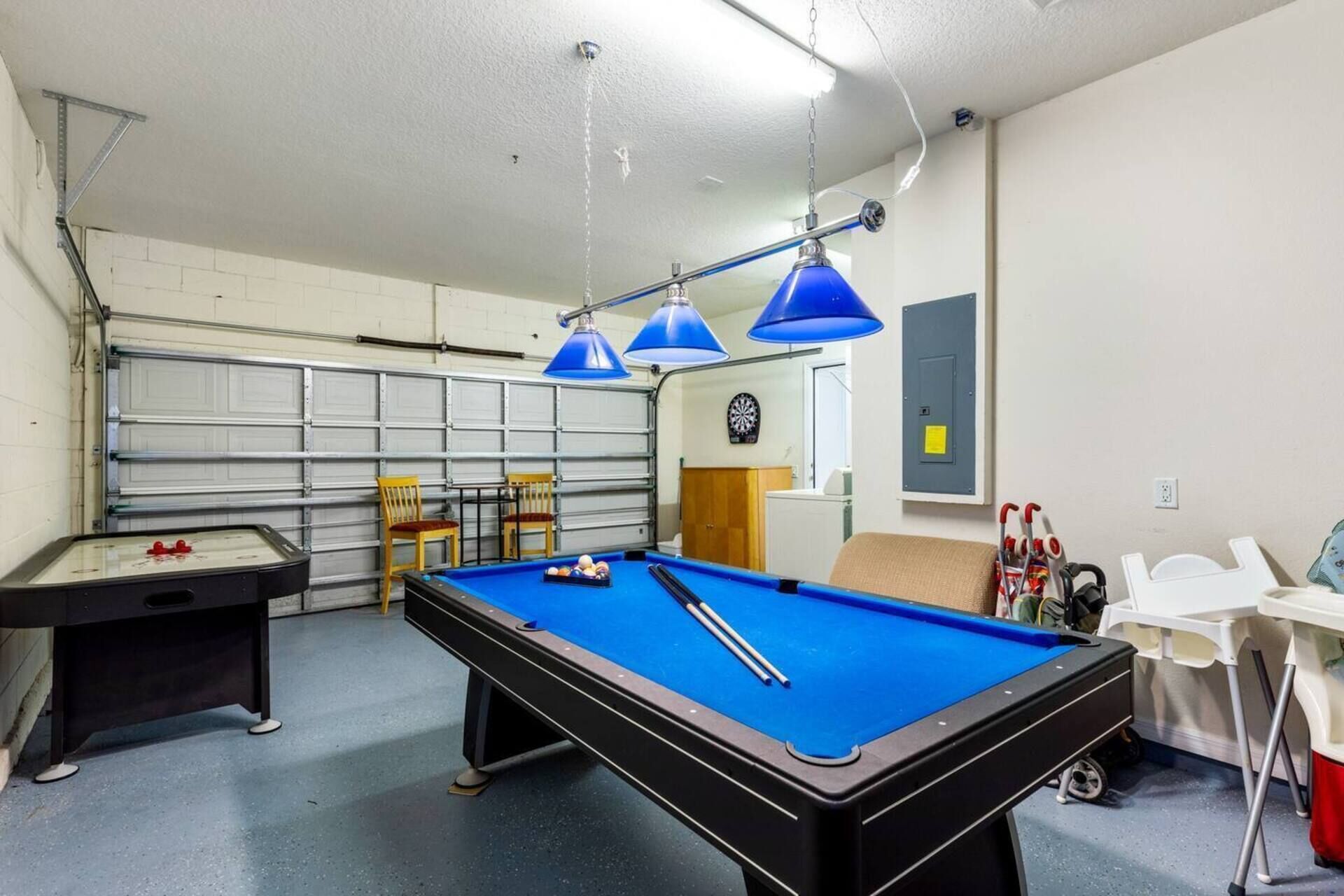 Een pooltafel met blauwe verlichting, een dartbord, en een kleine pooltafel in een ruimte met een garage deur en stoelen.