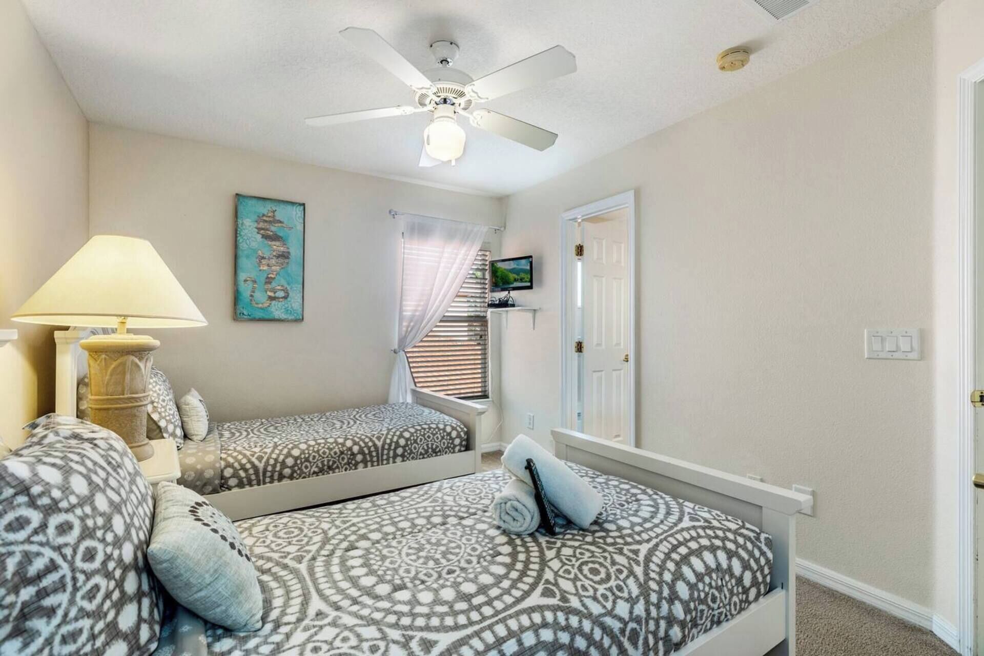 A bedroom with two beds, a ceiling fan, a lamp, a painting, and a window with blinds.