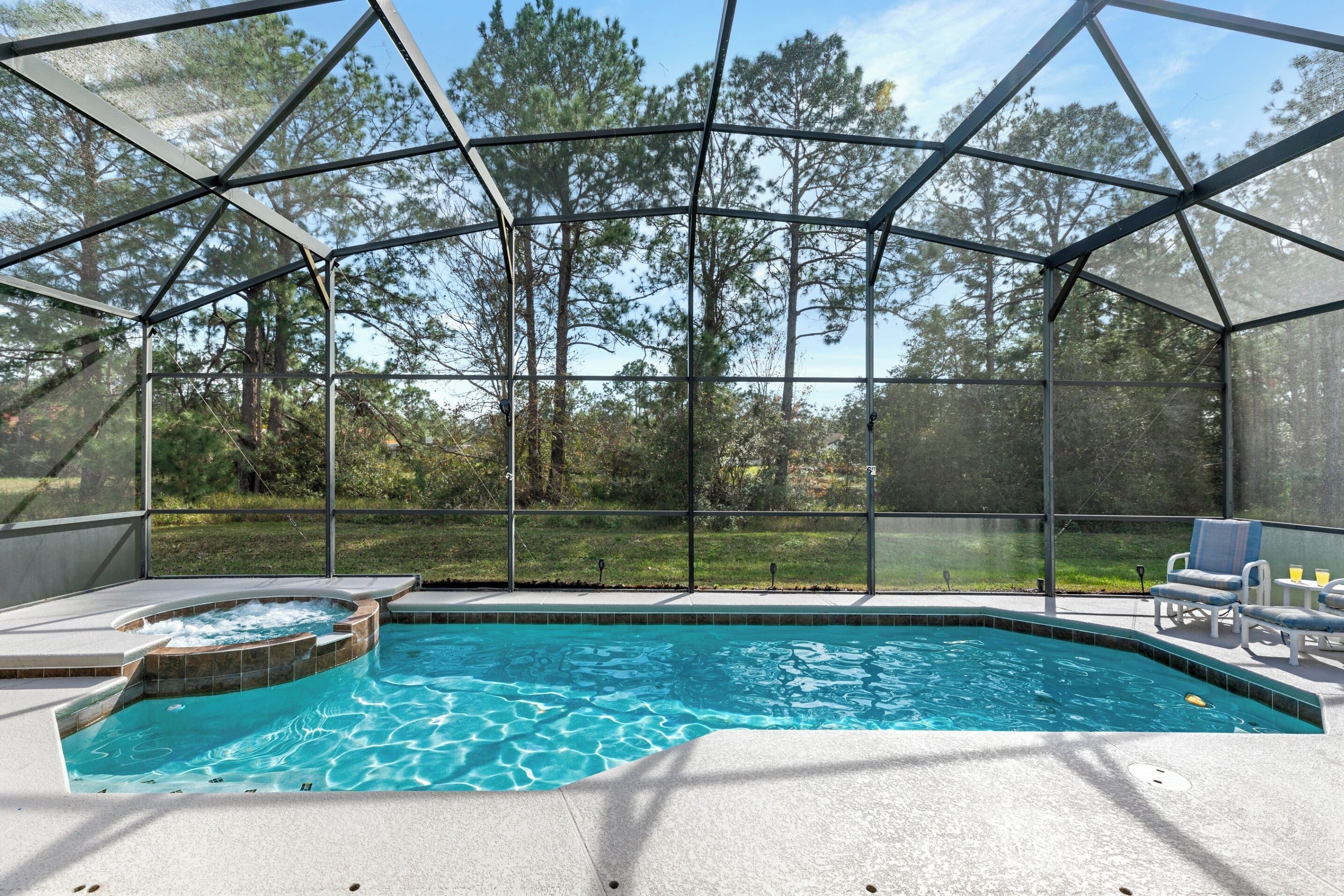 A swimming pool with a hot tub, surrounded by a covered area with chairs and a view of trees.