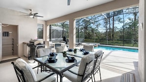 A spacious outdoor dining area with a glass table and chairs, a ceiling fan, and a view of a swimming pool and trees. - Gorgeous Davenport 5 Bed Villa With Pool Hot Tub (Davenport)