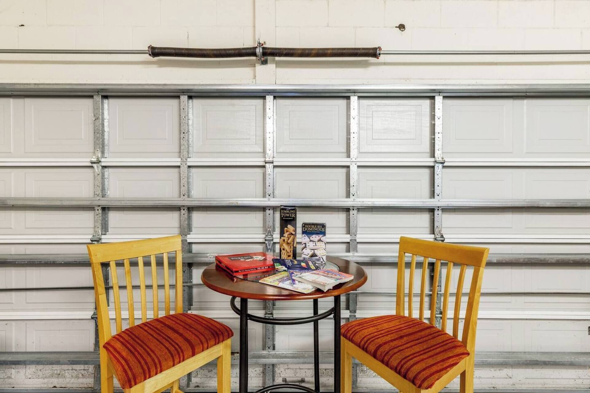 A garage with a closed white door, two wooden chairs with striped cushions, and a small round table with a box of dominoes and magazines.