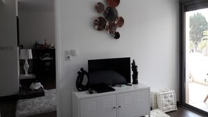Una sala de estar moderna con un televisor de pantalla plana sobre un mueble blanco, arte decorativo en la pared y vistas al exterior a través de un ventanal grande.