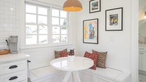 Une cuisine lumineuse avec des meubles blancs, une machine à café, une fenêtre offrant une belle vue, une table ronde blanche et un banc agrémenté de coussins à motifs.