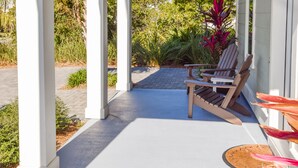 A covered porch with two wooden chairs, white columns, and a view of greenery and a house in the background. - Sea Glass Cottage ~ Private Pool ~ Private Beach Access ~ Golf Cart Included! (Miramar Beach)