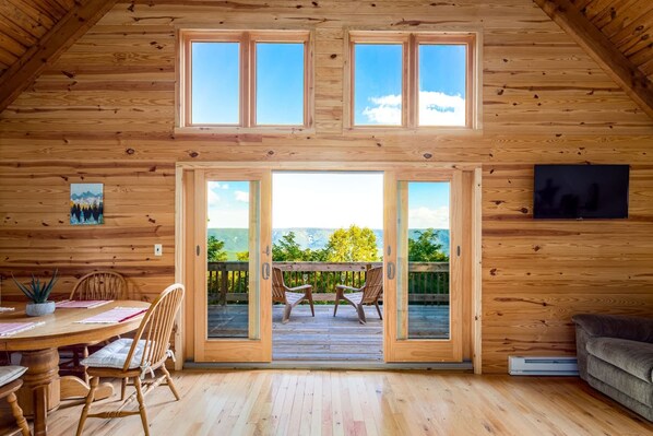 A wooden cabin interior with a dining area, a sofa, and a balcony with a view of the mountains.