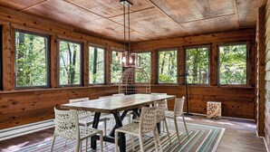 A wooden cabin interior with a large dining table, white chairs, a hanging light fixture, and large windows offering a forest view. - Huge Cabin w Hot Tub, Deck, Grill & WiFi (Berkeley Springs)