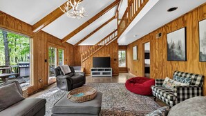 A spacious living room with a high ceiling, wooden beams, a large TV, and a cozy seating area with a red bean bag. - Huge Cabin w Hot Tub, Deck, Grill & WiFi (Berkeley Springs)