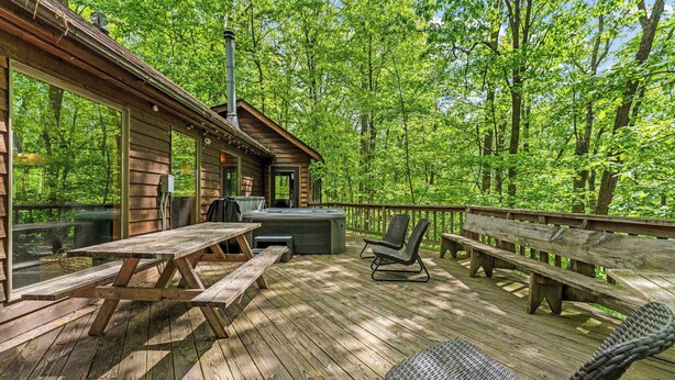 A wooden deck with picnic tables and chairs, a hot tub, and a view of a forest. - Huge Cabin w Hot Tub, Deck, Grill & WiFi (Berkeley Springs)