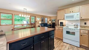 A modern kitchen with wooden cabinets, a large island, and a dining area with a table and chairs. - Secluded Oasis w Hot Tub, Screened-in Porch, WiFi! (Berkeley Springs)