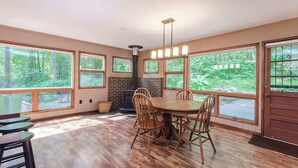 A spacious dining area with a large wooden table and chairs, surrounded by large windows offering a view of the forest. - Secluded Oasis w Hot Tub, Screened-in Porch, WiFi! (Berkeley Springs)