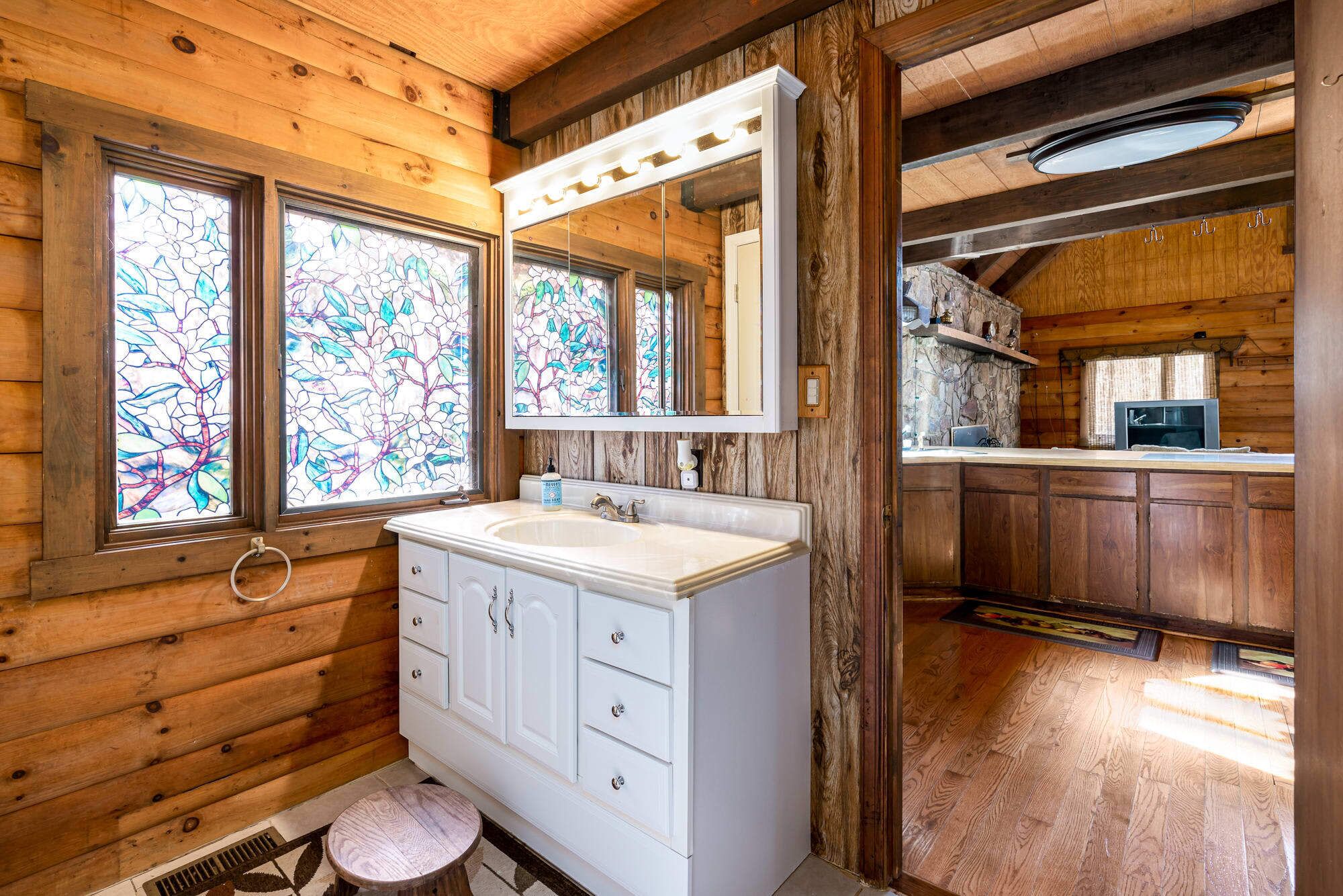 Baño de cabaña de madera con tocador blanco, un espejo grande y ventanas de vitral.