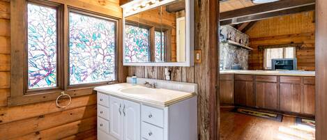 Baño de cabaña de madera con tocador blanco, un espejo grande y ventanas de vitral.