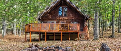 A cabin with a deck, surrounded by trees and a fire pit.