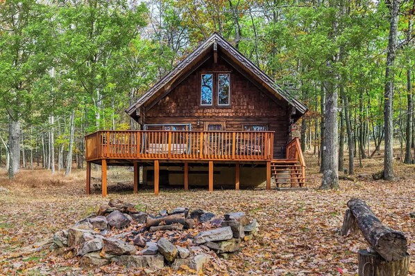 A cabin with a deck, surrounded by trees and a fire pit.