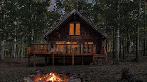 A cabin with a deck, surrounded by trees, and a fire pit in front.